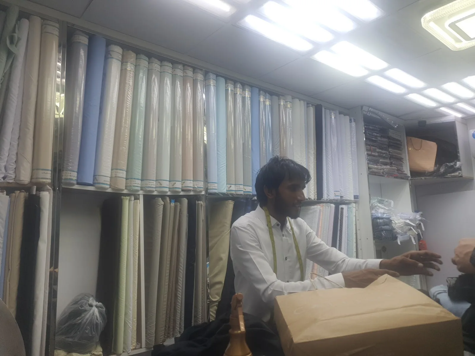 Rolls of suiting and shirting fabric stacked on shelves with a tailor