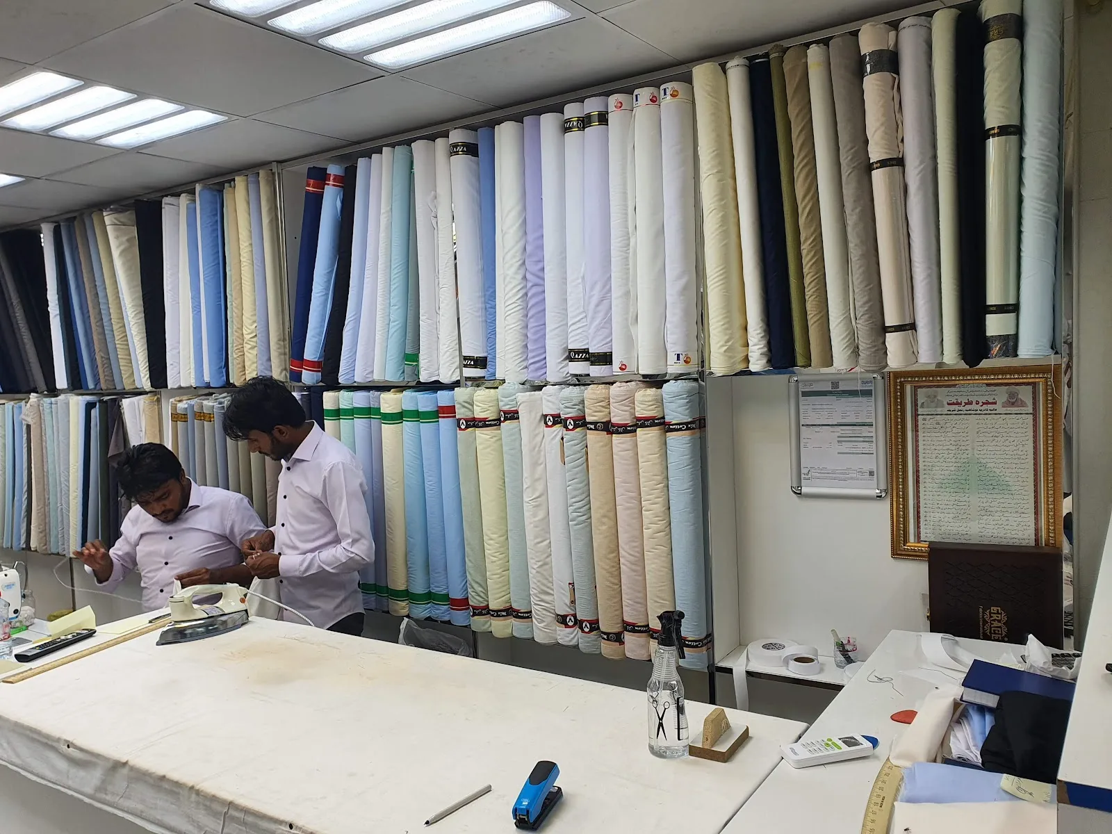 Tailors pressing and finishing garments beside a wall of rolled cloth