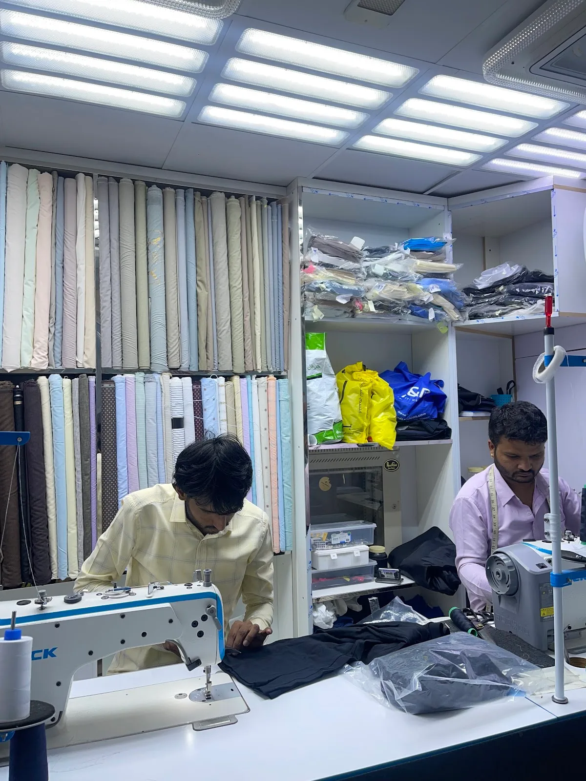 Two tailors stitching at sewing machines in front of a wall of fabric