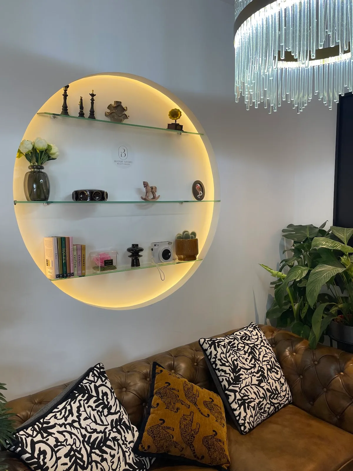 Blonde Studio lounge with a backlit circular display shelf, chesterfield sofa, plants and a glass-rod chandelier
