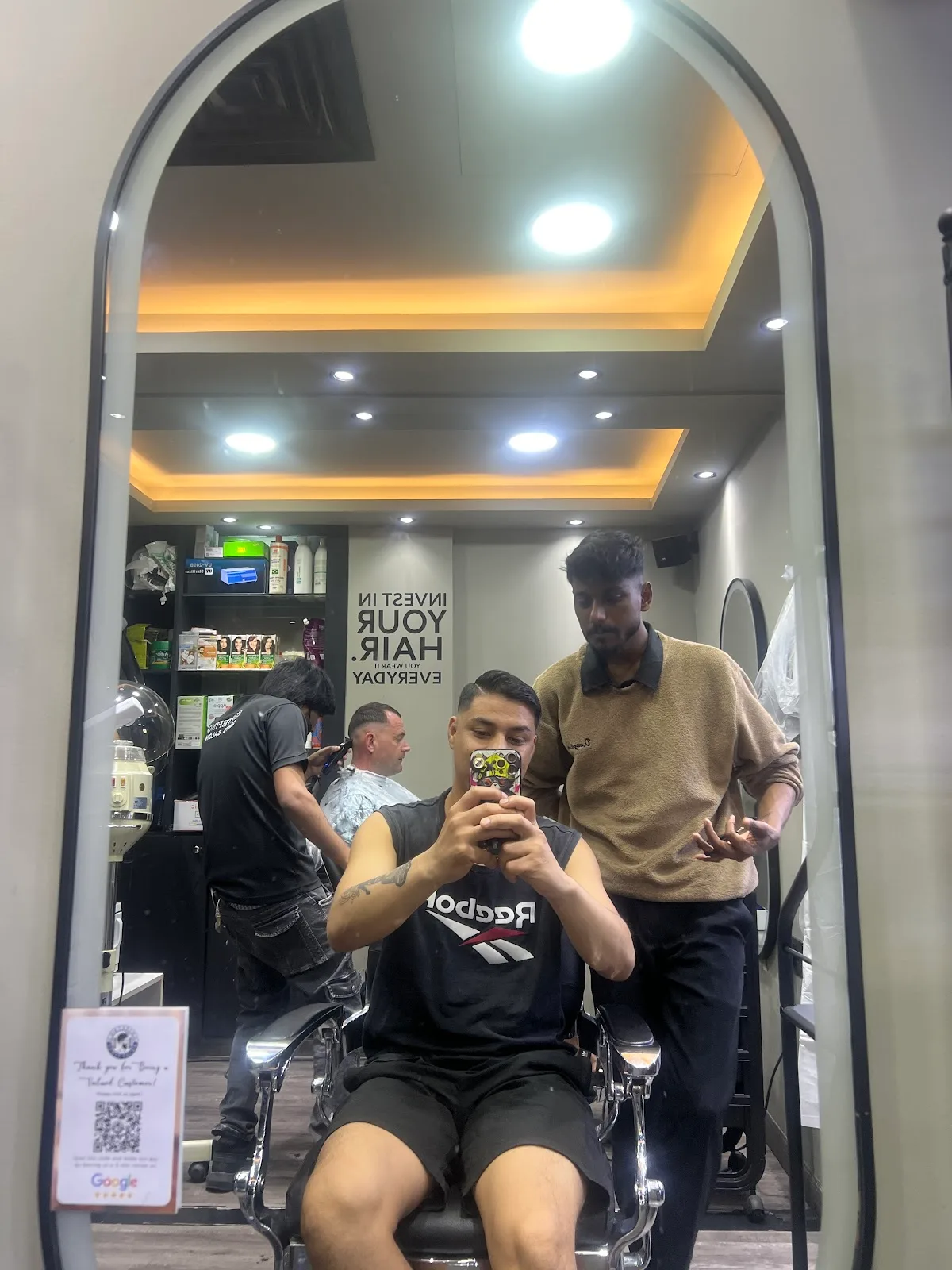 A customer in the barber chair reflected in CorteFino's arched mirror with the wall slogan invest in your hair