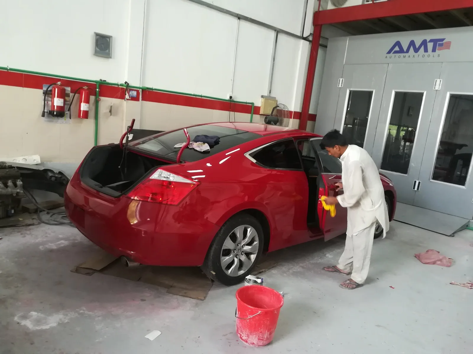 Technician detailing a red coupe beside the spray paint booth at Muhammad Asghar Garage