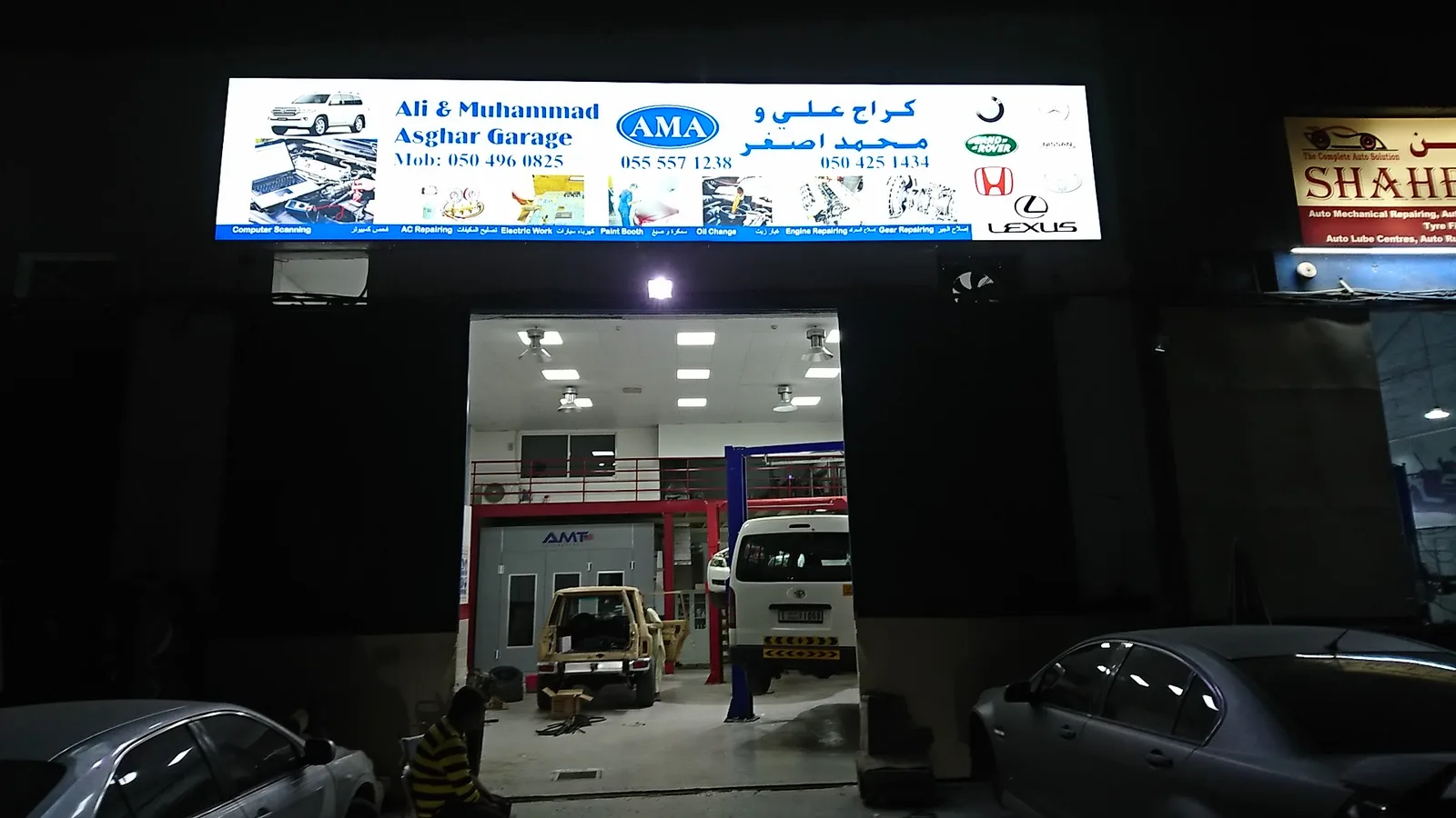 Lit signboard of Ali & Muhammad Asghar Garage at night in Umm Ramool, Dubai, with cars inside the workshop