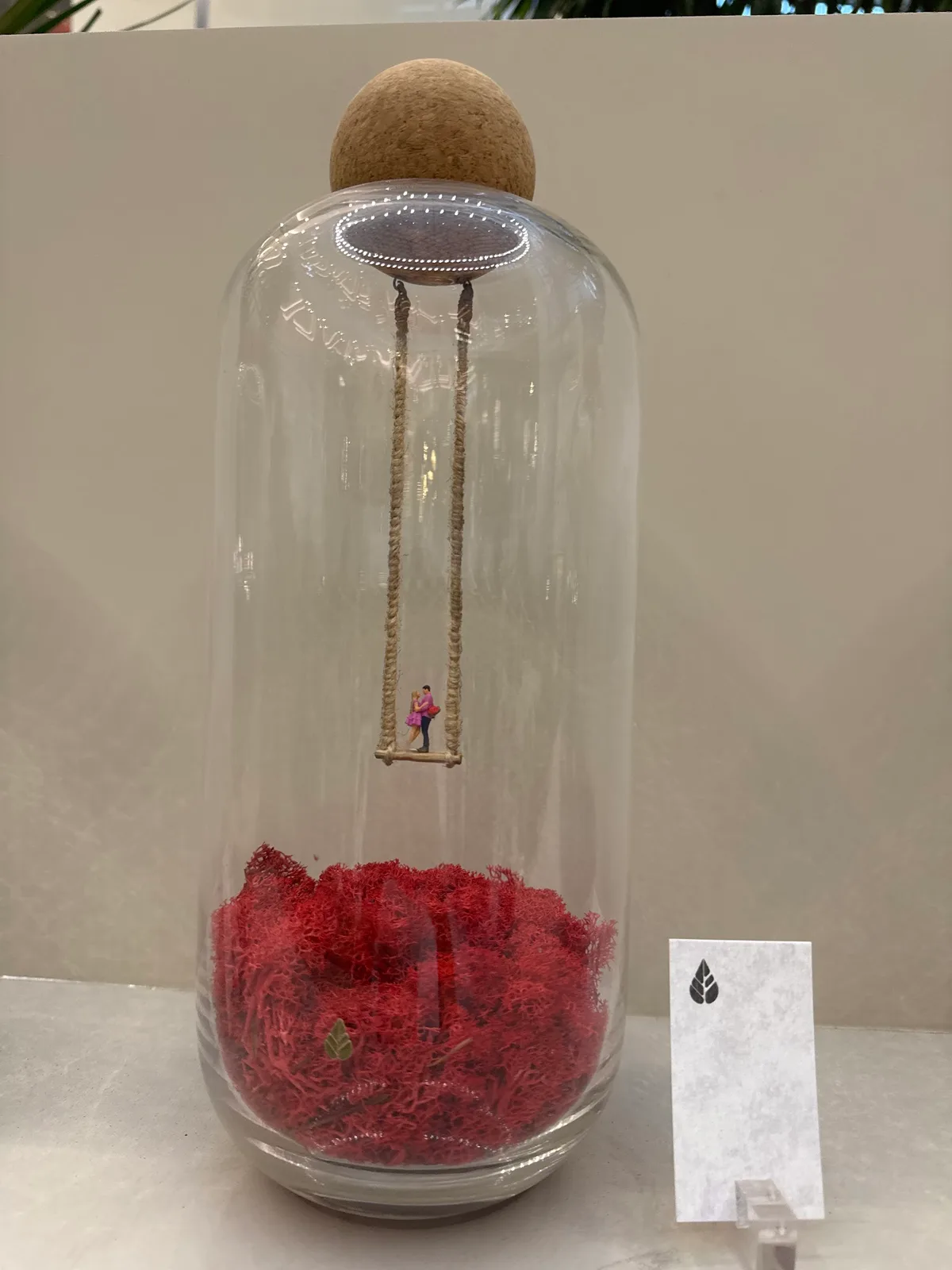 Glass bottle terrarium with a tiny couple on a swing above red preserved moss at Folivana