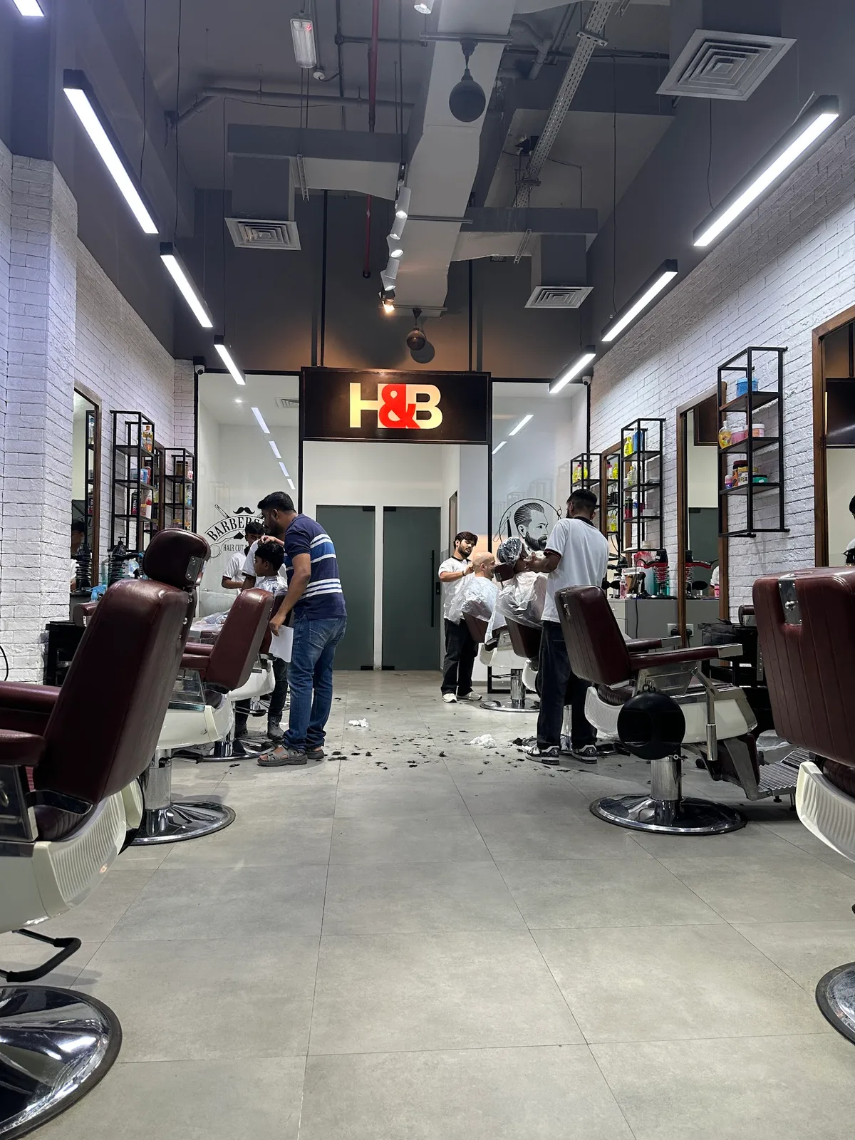 Inside H&B Gents Salon, Discovery Gardens — barbers at work beneath the lit H&B sign