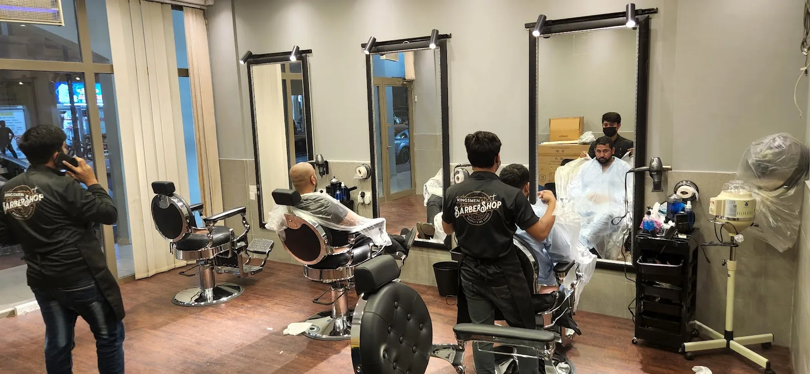 Inside Kingsmen Barbershop — barbers in branded shirts working at the chairs by tall mirrors