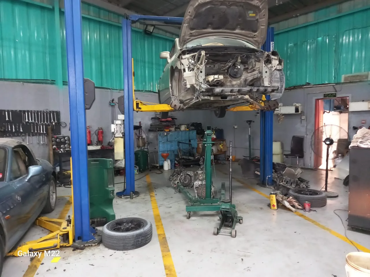 A vehicle raised on a two-post lift with the front end stripped and an engine and gearbox out on a stand below.