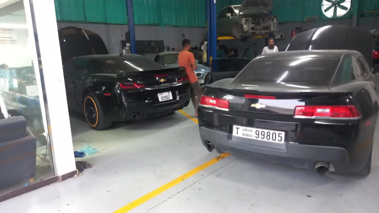 Cars including a black and a grey Chevrolet Camaro inside the Al Quoz workshop, with mechanics at work and a vehicle on the lift in the background.