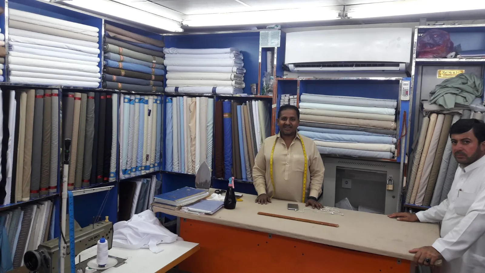 Tailor with a measuring tape around his neck standing at the counter inside Qasar Al Ajaz, fabric stacked on the walls behind