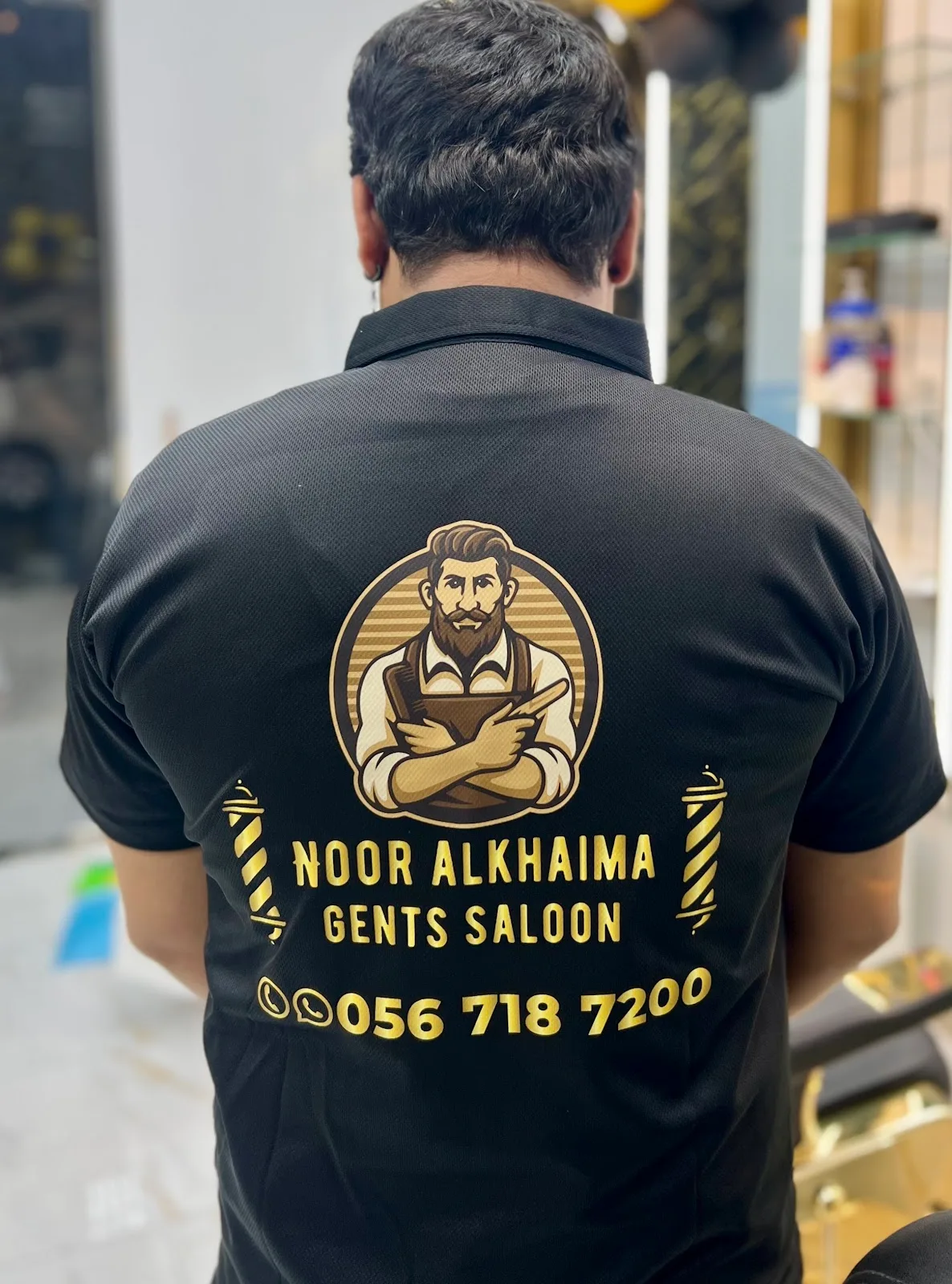 A Noor Al Khaima team member in the salon's black polo shirt, printed with the gold barber emblem and the phone number 056 718 7200