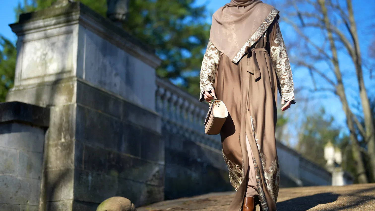 Model in a hand-embroidered brown abaya carrying a handbag outdoors